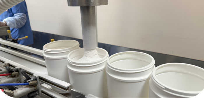 White supplement containers being filled with powder on a production line, with a worker and industrial equipment visible in the background.